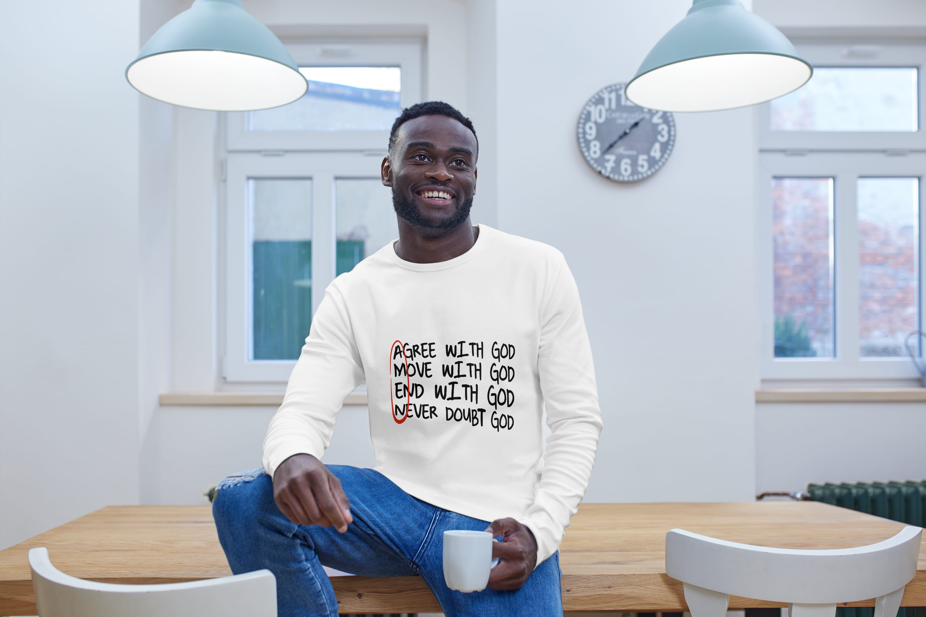 Man sitting at a table wearing a white sweater with text Agree with God Move with God End with God Never Doubt God, smiling in a room with large windows.