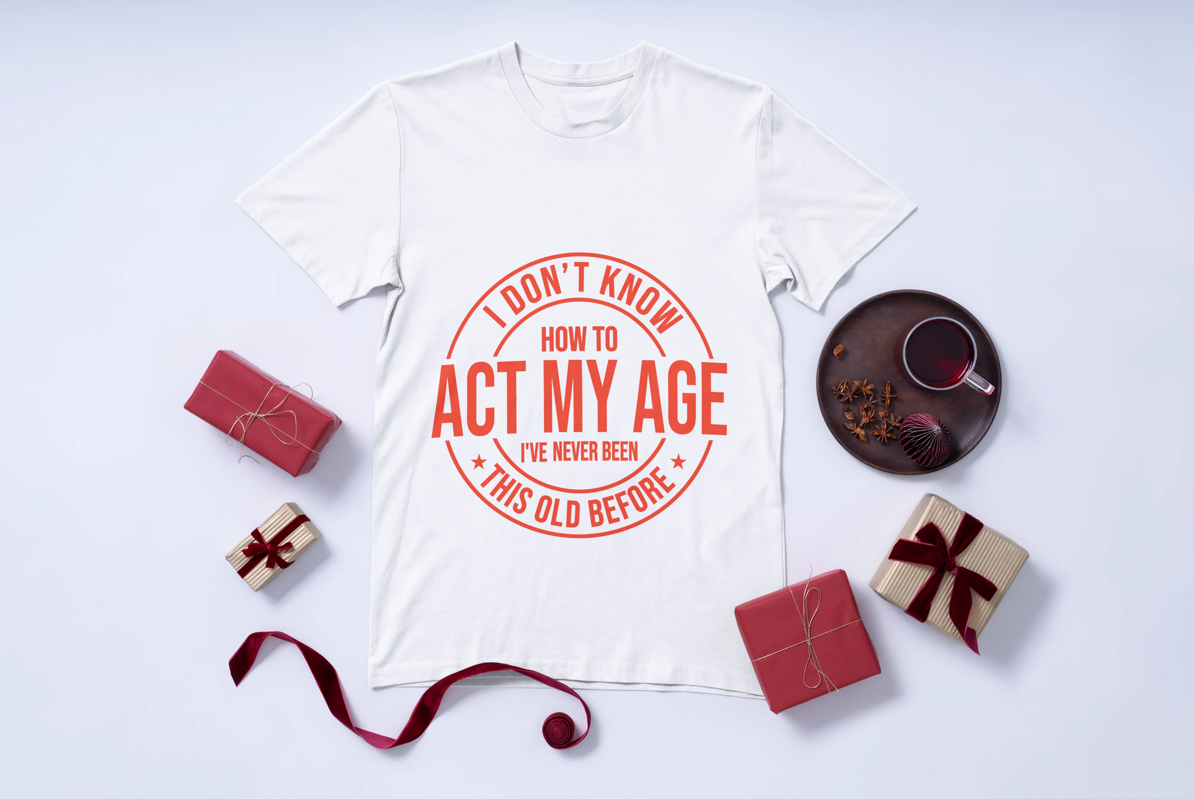 White t-shirt with red text I don't know how to act my age I have never been this old before on a white background with small gifts and a cup