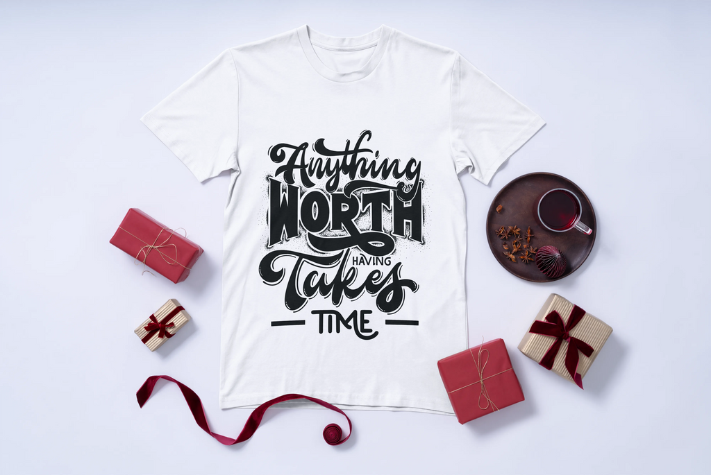 White t-shirt with black text 'Anything Worth Having Takes Time' on a light gray background with decorative items.