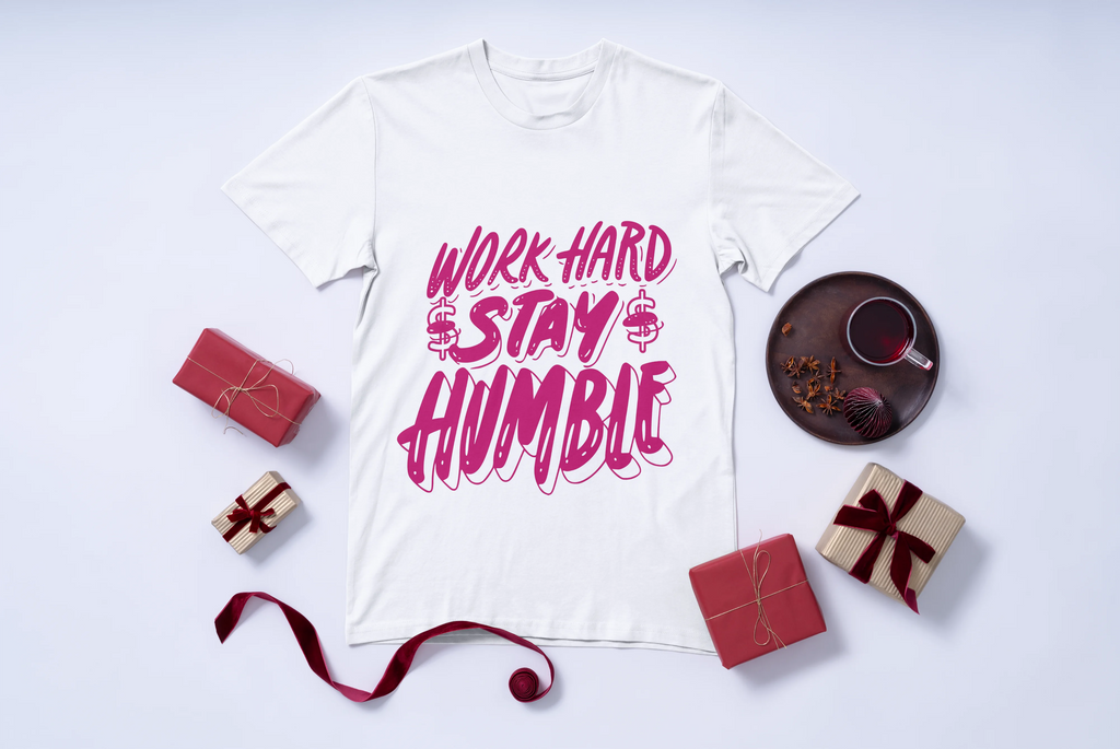 White t-shirt with 'Work Hard Stay Humble' text in pink color on a white background with decorative items.